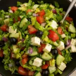 Mediterranean Greek salad recipe with fresh romaine lettuce, cucumbers, tomatoes, black olives, red onions, and feta cheese cubes in a black bowl