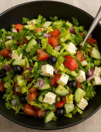 Mediterranean Greek Salad Recipe Mediterranean Greek salad recipe with fresh romaine lettuce, cucumbers, tomatoes, black olives, red onions, and feta cheese cubes in a black bowl