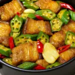 Stir fried pork belly recipe with green okra, red chili slices, and whole garlic cloves in a savory sauce, presented in a black serving bowl.