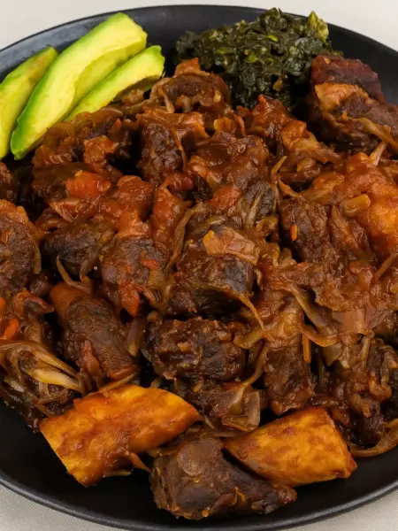 Best Ugandan Pork Stir Fry (Kikalayi Pork) Ugandan-style pork stir fry, also known as Kikalayi pork, served with matooke, avocado slices, and cabbage on a black tray.