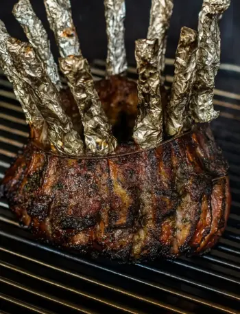 Best Crown Roast of Lamb for Festive Holidays Juicy, deeply browned crown roast of lamb cooking on a grill or oven rack, with the top of each rib bone covered in crinkled silver foil, perfect for a family dinner, Easter party, Thanksgiving, or Christmas dinner.