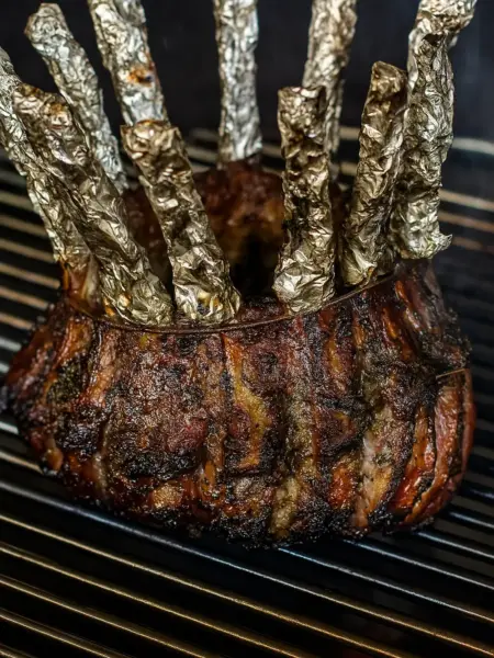 Best Crown Roast of Lamb for Festive Holidays Juicy, deeply browned crown roast of lamb cooking on a grill or oven rack, with the top of each rib bone covered in crinkled silver foil, perfect for a family dinner, Easter party, Thanksgiving, or Christmas dinner.
